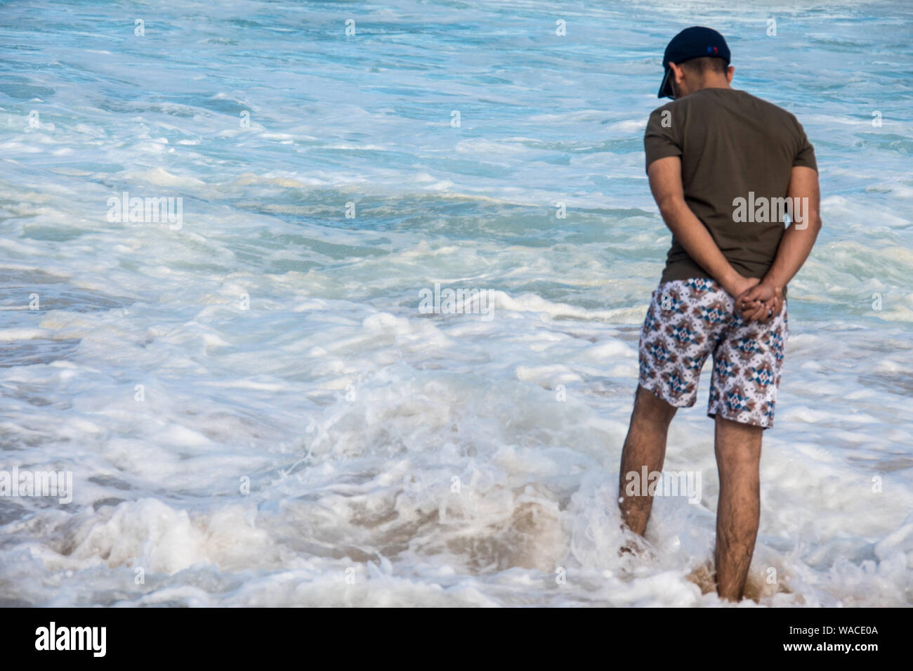 Ein Mann in lässige Beachwear nachdenklich im Ozean stehen mit den Händen hinter seinem Rücken, während sie tief in Gedanken. Die Wellen plätschern auf ihn Stockfoto