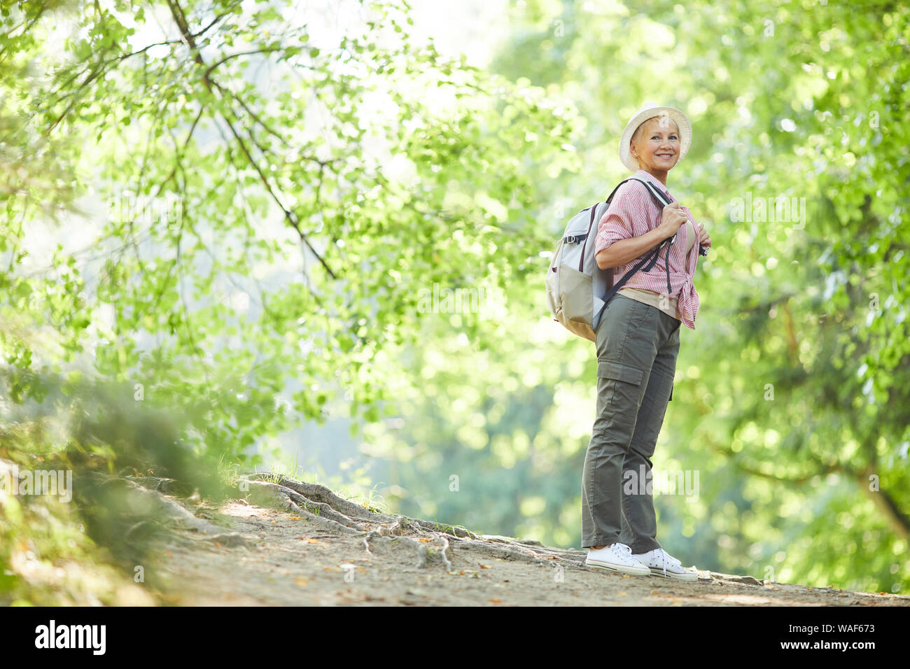 Reife Frau in Freizeitkleidung mit Rucksack auf dem Rücken stehen und Lächeln bei Ihrem Spaziergang in den Wald im Sommer Stockfoto