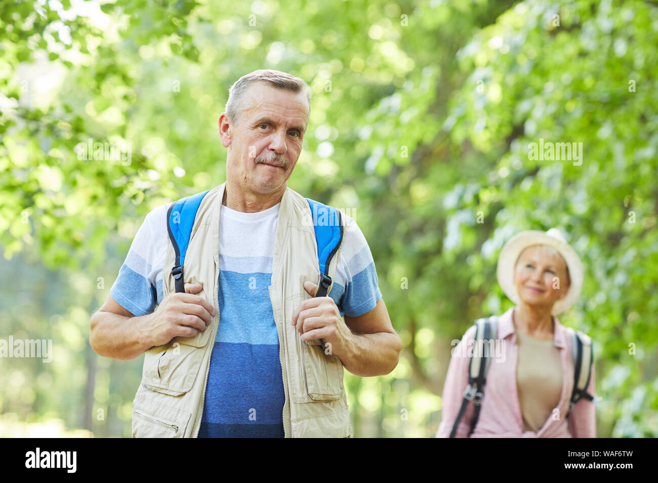 Portrait von älteren Mann in lässige Kleidung und mit Rucksack an der Kamera, während eine Reise in den Wald mit Frau im Hintergrund Stockfoto
