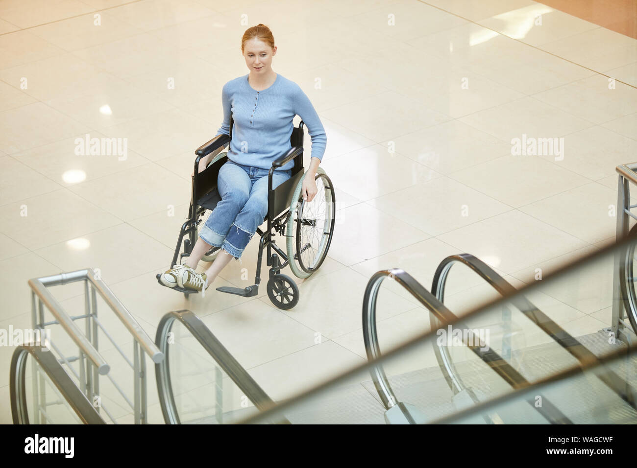 Behinderte Frau in Freizeitkleidung sitzt im Rollstuhl und Rolltreppe in modernes Gebäude Stockfoto