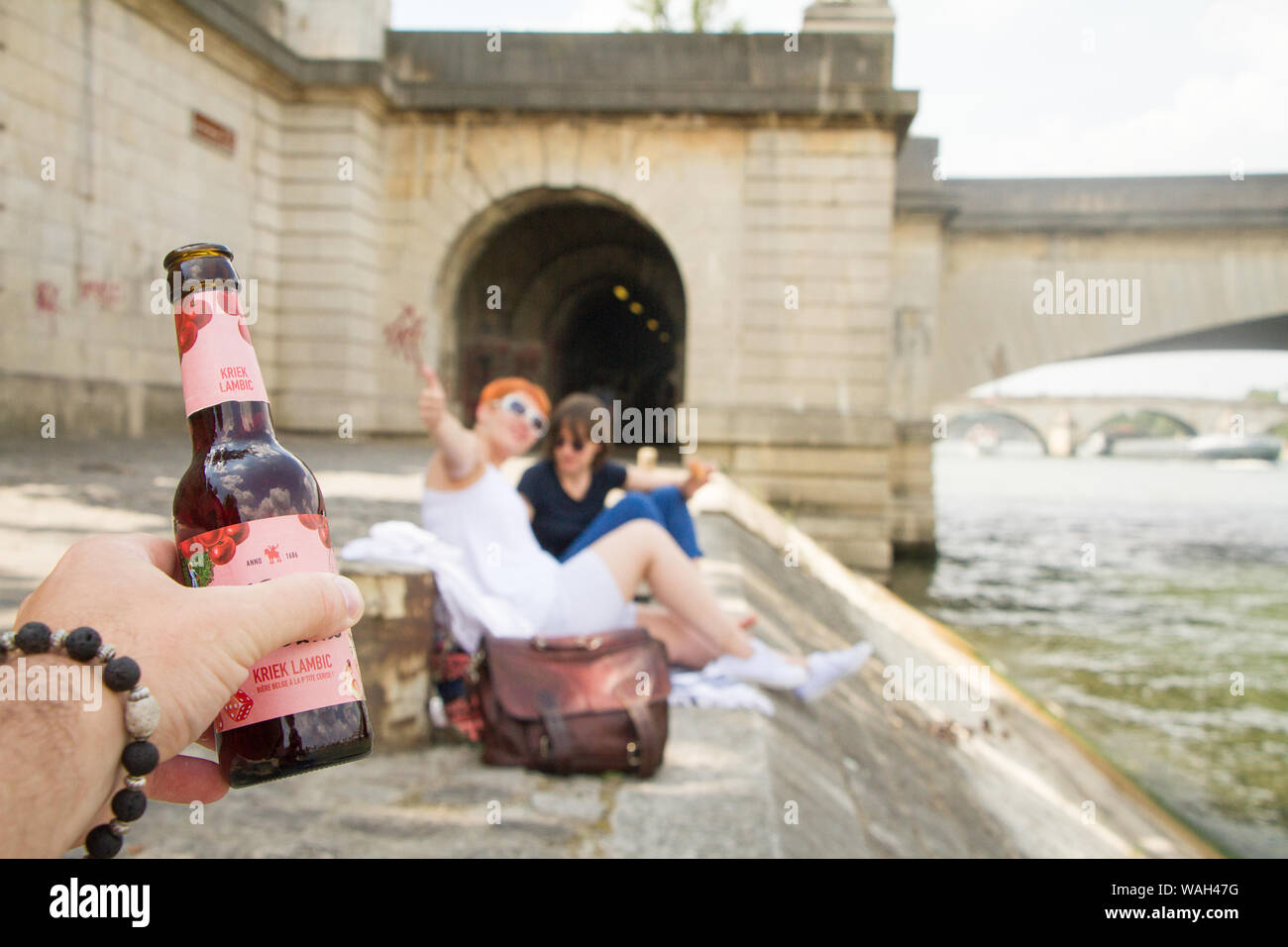 Paris, Frankreich, 07.Juli 2018: Eine Hand hält eine Flasche Bier auf einen unscharfen Hintergrund einer Picknick von zwei Mädchen auf der Seine Damm Stockfoto