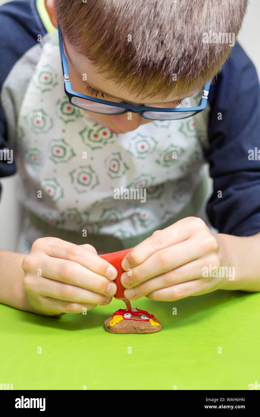 Der junge schmückt ein Zimt Cookie. Er hält eine Tube rot Vereisung in seine Hände und drückt es auf dem Kuchen. Stockfoto