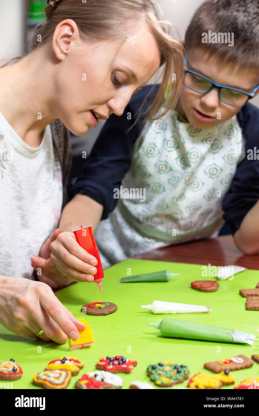 Mutter und Sohn in der Küche. Eine Frau schmückt ein Zimt Kuchen mit einer tube rot Vereisung. Der Sohn beobachtet aufmerksam, was seine Mutter tut. De Stockfoto