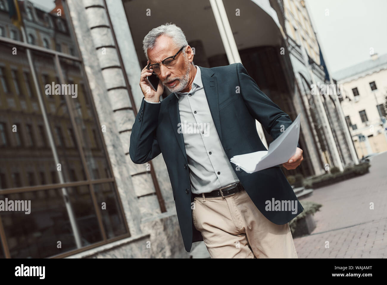 Senior Geschäftsmann in Anzug holding Papiere und Unterhaltung am Telefon beim Gehen im Freien. Wirtschaft, Technologie, Kommunikation und Personen Konzept. Horizontale erschossen. Stockfoto
