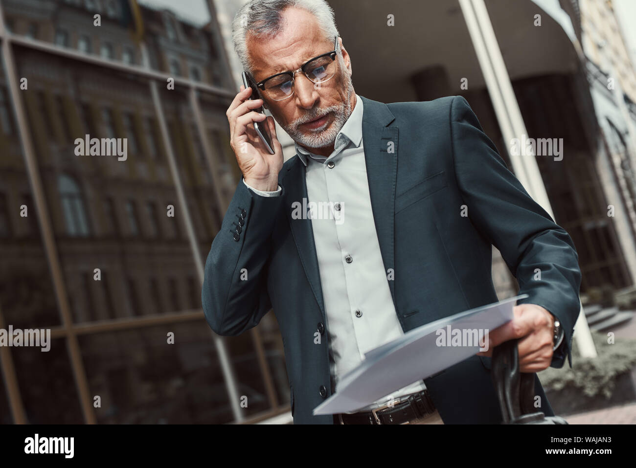 Senior Geschäftsmann in Anzug holding Papiere und Unterhaltung am Telefon beim Gehen im Freien. Wirtschaft, Technologie, Kommunikation und Personen Konzept. Horizontale erschossen. Stockfoto