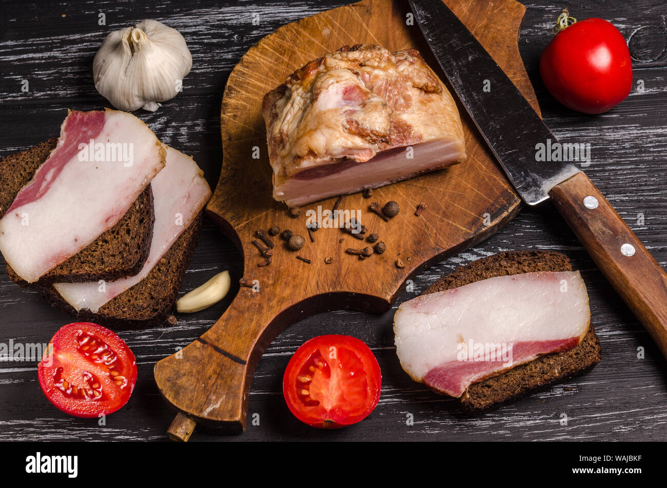 Saftige geräucherter Speck mit Sandwiches, Gewürze und rohes Gemüse auf einem schwarzen Holztisch. Stockfoto