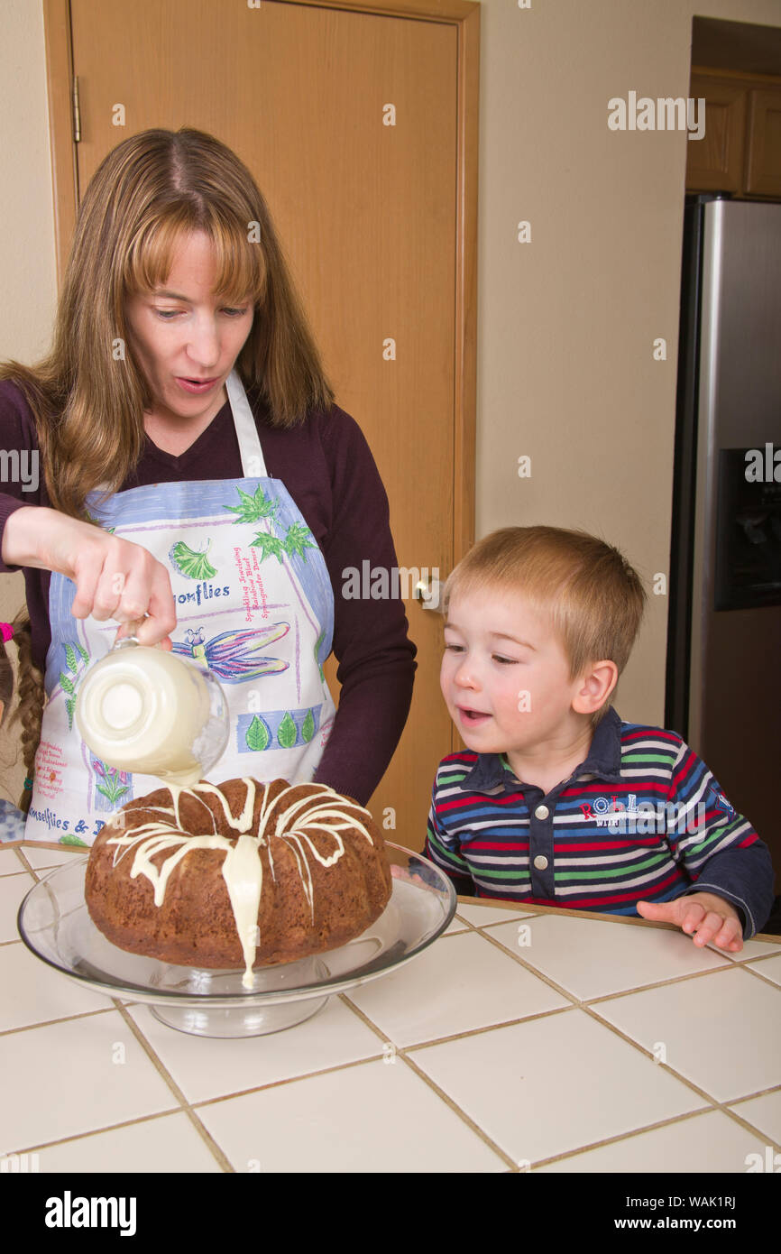 Mutter gießen weiße Schokolade auf Schokolade bundt Kuchen Glasur während drei Jahre alten Sohn Uhren. (MR, PR) Stockfoto