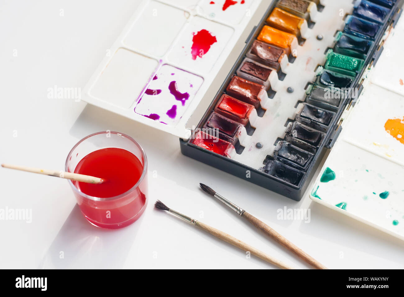 Aquarell Palette mit viele Schatten, ein paar flauschige Bürsten und ein Glas Schale Wasser für Waschbürsten, bereits rot eingefärbt. Stockfoto