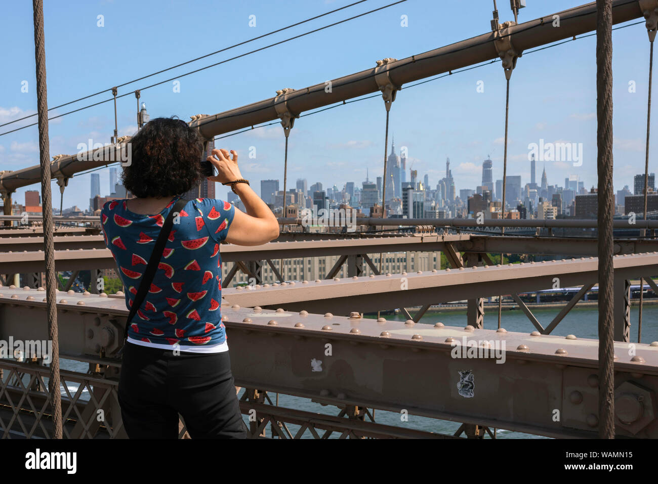 New York Tourist, Ansicht von hinten im Sommer eine alleinreisende weibliche Touristen stehen auf der Brooklyn Bridge und nehmen ein Foto von Midtown Manhattan, New York City, USA Stockfoto