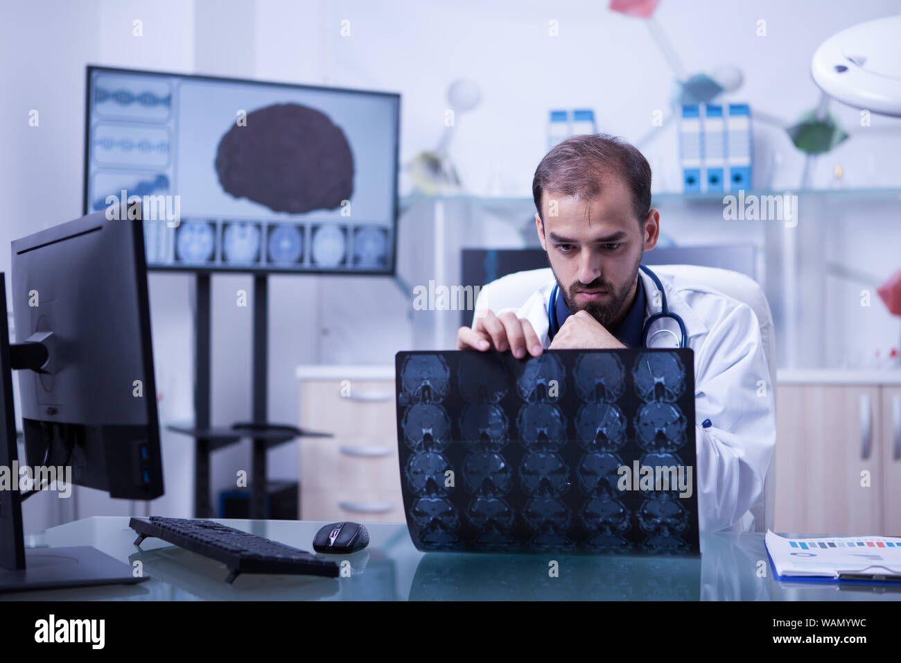 Schöne bärtige Doktor an ein Gehirn Radiographie. Arzt sitzt an seinem Schreibtisch in einem Schrank Kontrolle eine Gehirn-CT-Scan. Stockfoto
