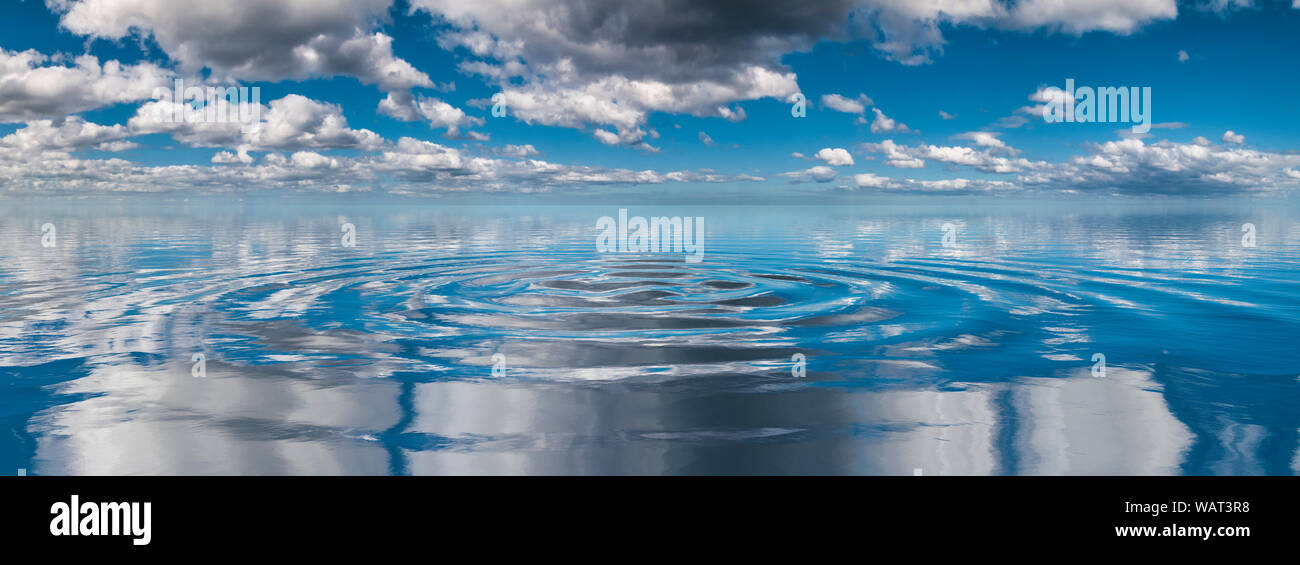 Kreisförmige Wellen breiten sich über ruhiges Wasser aus und reflektieren einen bewölkten Himmel mit verstreuten Kumuluswolken, wodurch eine friedliche, symmetrische Szene entsteht Stockfoto
