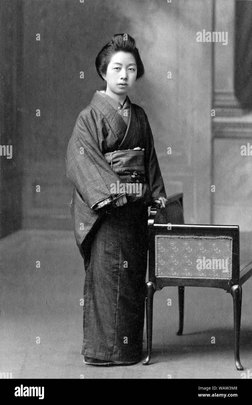 [1910s Japan - Japanische Frau im Kimono] - junge japanische Frau im Kimono steht neben einem Stuhl. 20. Jahrhundert vintage Silbergelatineabzug. Stockfoto