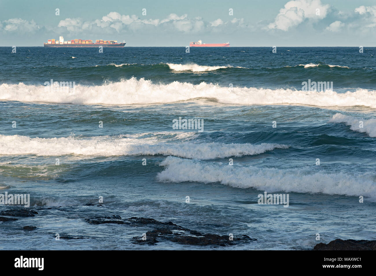 Containerschiffe auf dem Ozean mit dem Brechen der Wellen und Felsen im Vordergrund. Stockfoto