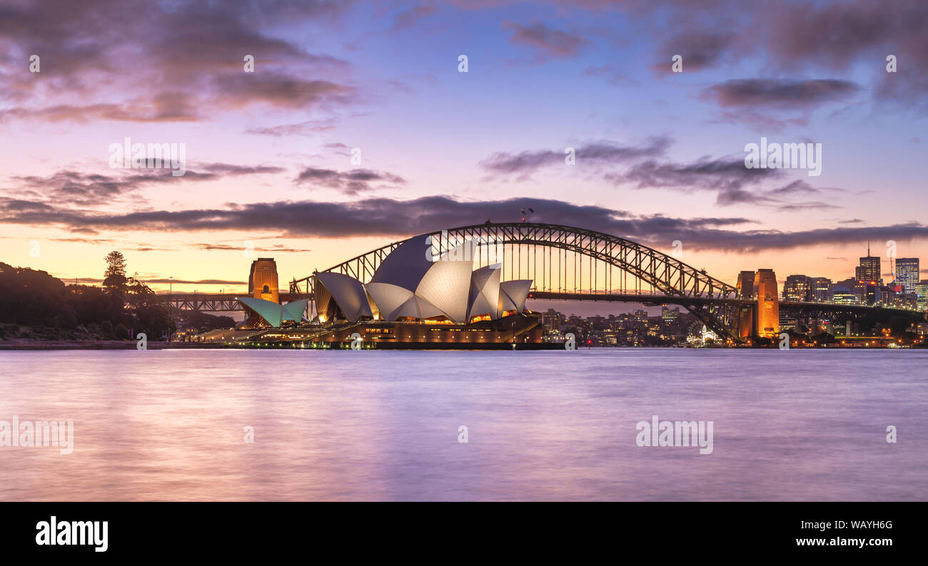 Sydney Opera House und die Brücke in der Abenddämmerung. Sydney Skyline Panorama mit bunten lila Reflexion aus der Distanz. Sonnenuntergang, NSW, Australien. Stockfoto