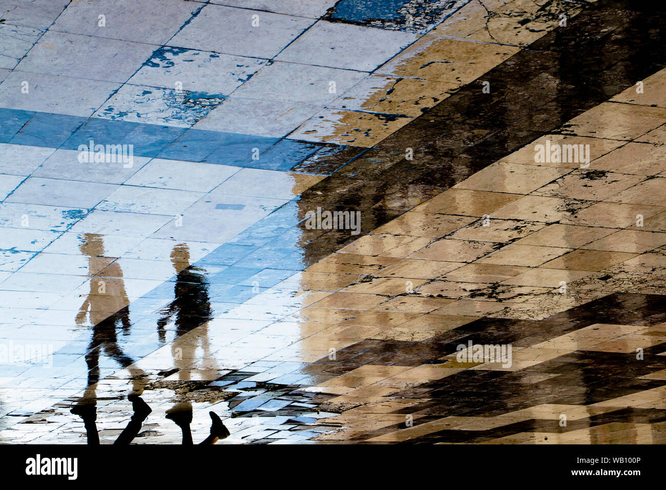 Unscharfe Reflexion Schatten Silhouetten der beiden jungen Männer gehen auf eine vintage Pflaster auf nasser Straße der Stadt, einer trägt eine Flasche und einen Sac Stockfoto