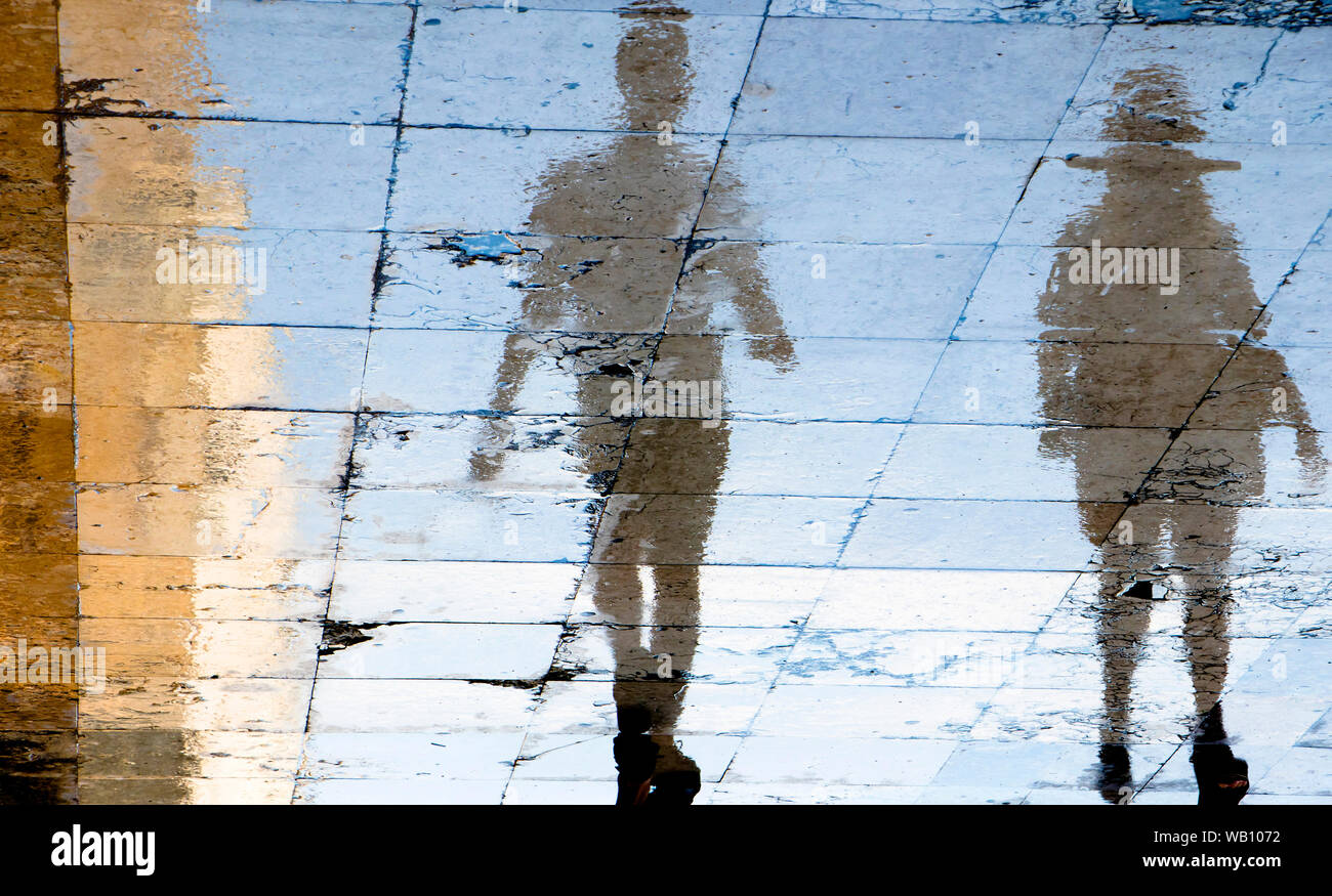 Unscharfe Reflexion Schatten Silhouetten von zwei Menschen zu Fuß auf einer nassen Straße an einem Sommertag in der Stadt Stockfoto