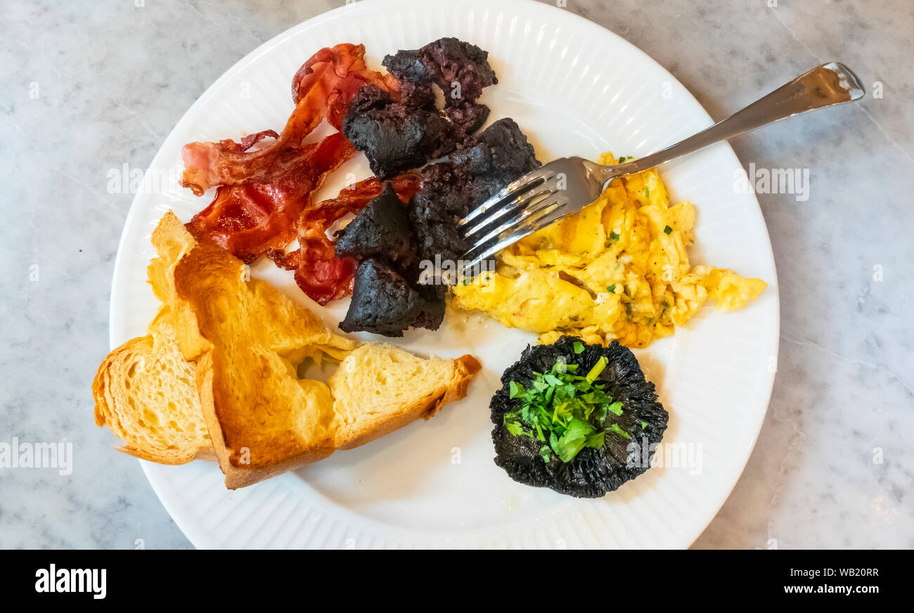 Klassische heiße französisches Frühstück mit Rührei, Champignons, Speck, Boudin Noir, und Toast. Stockfoto