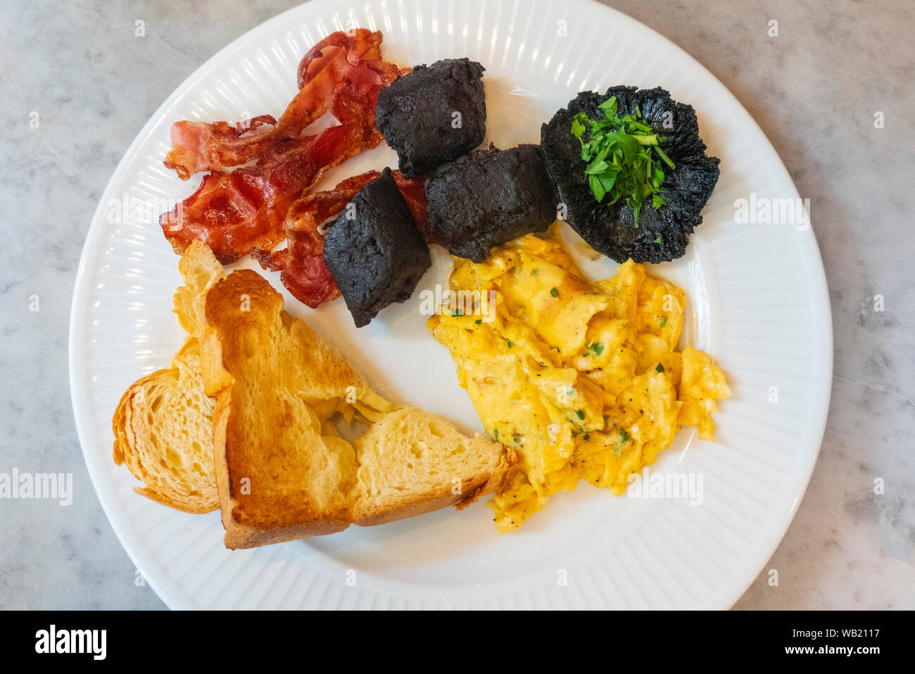 Klassische heiße französisches Frühstück mit Rührei, Champignons, Speck, Boudin Noir, und Toast. Stockfoto
