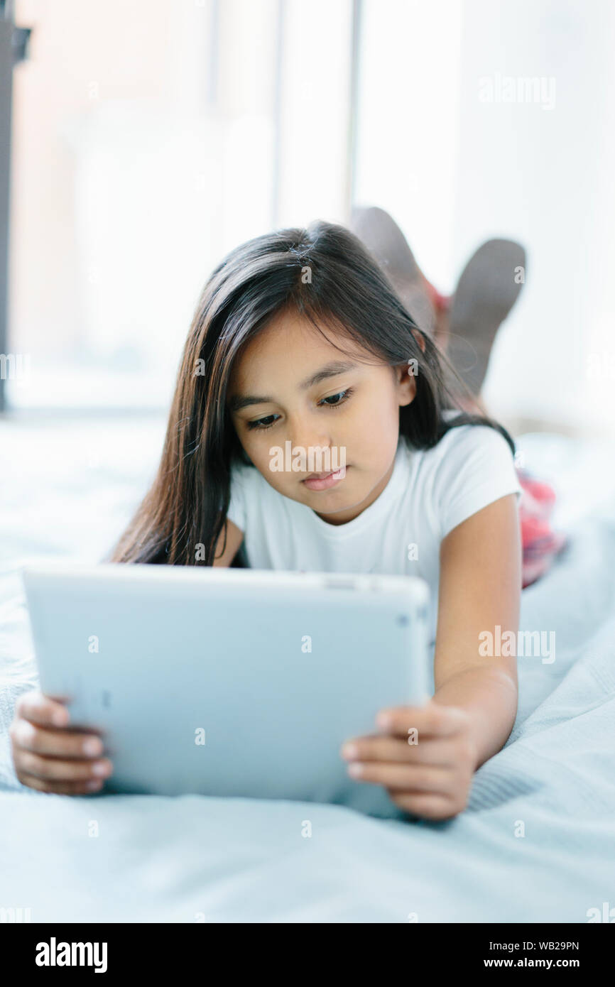 Ein 9-jähriges Mädchen liegt auf ihrem Bett. Sie ist auf der Suche an Ihrem  Tablet-PC. Sie ist für die Schule angezogen. Es ist tagsüber. Sie hat  dunkle lange Haare und Hispanic wurde