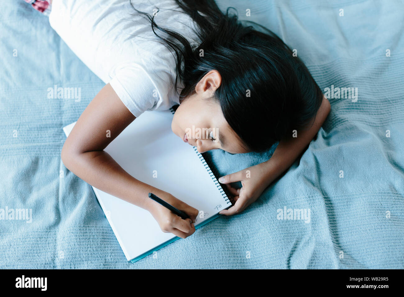 Ein 9-jähriges Mädchen liegt auf einer Decke auf dem Bett. Sie ist schriftlich auf einem Notebook. Sie hat lange dunkle Haare. Sie hat Hispanic Ethnizität Features. Stockfoto