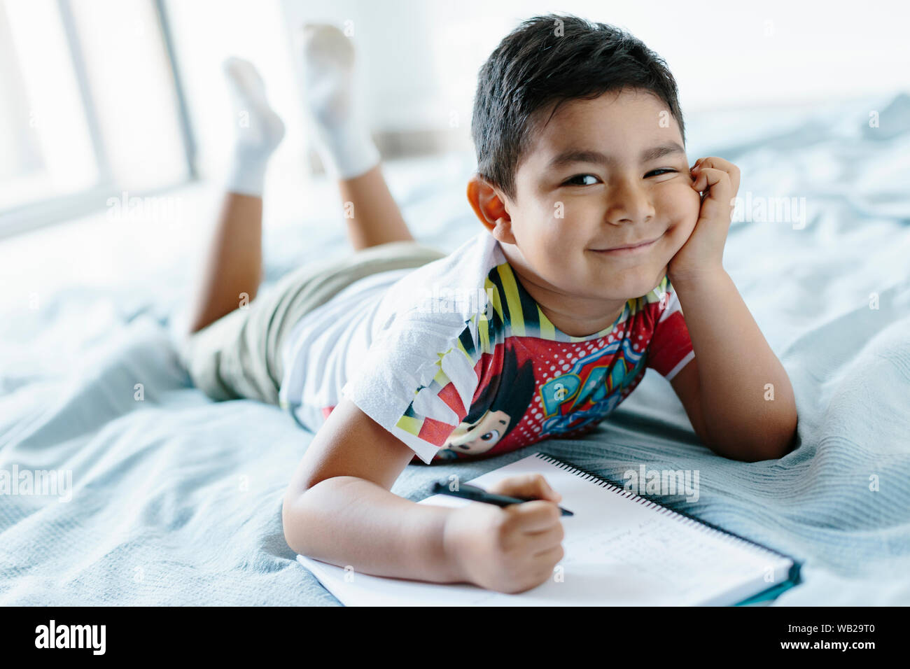 Ein 5-jähriger Junge liegt auf einer Decke auf dem Bett. Er sieht in die Kamera. Er hat Hispanic Ethnizität Features. Er lächelt. Er hat eine freche oder Art e Stockfoto