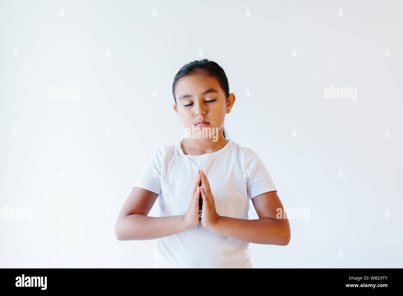 Ein 9-jähriges Mädchen sitzt ihr Namaste darstellen. Sie trägt weiße Kleidung. Sie hat Hispanic Ethnizität Features. Ihre Augen sind geschlossen. Stockfoto