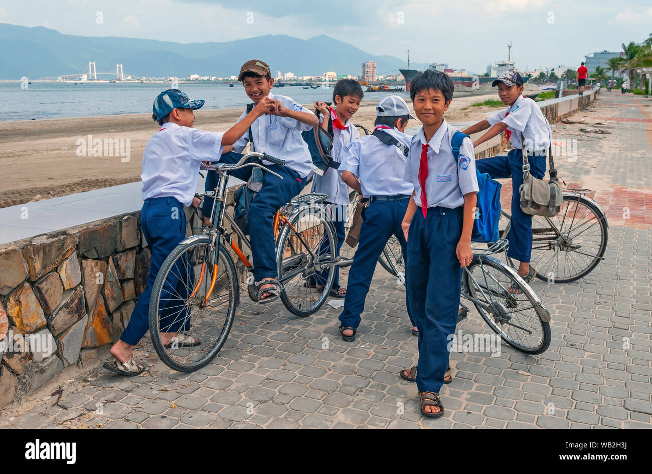 Gruppe von Jungen in Uniform mit Fahrrädern Spaß an der Uferpromenade von Danang mit seiner modernen Brücke, Central Vietnam. Stockfoto