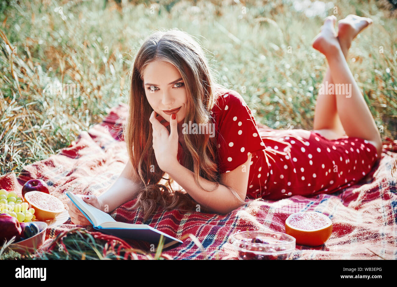 Picknick Szene: Süß lächelnde Mädchen liegen auf einem karierten und lesen Buch in Grove. Outdoor Portrait von hübsche junge Frau im roten Kleid Stockfoto