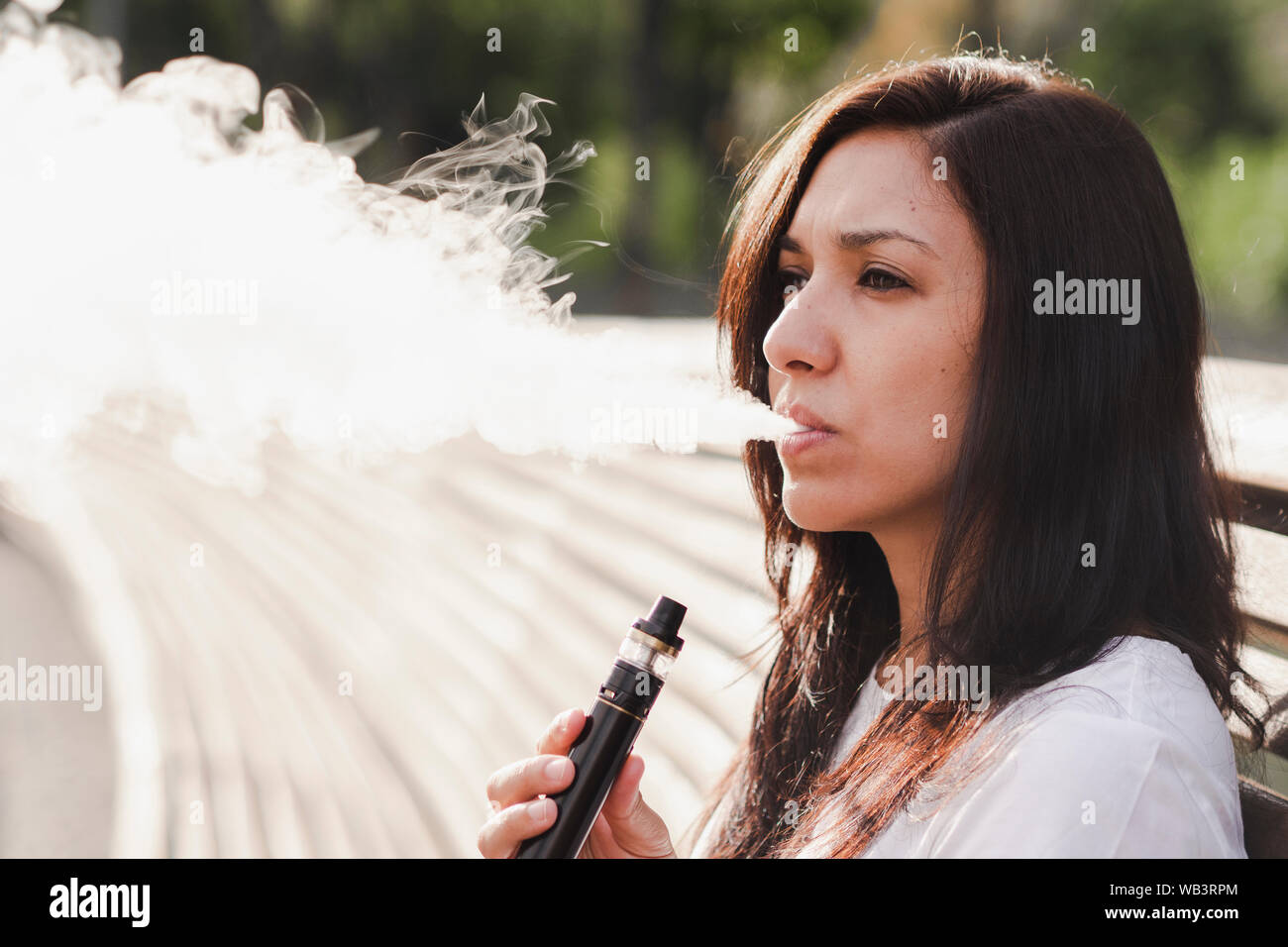 Junge Frau sitzt auf einer Parkbank rauchen mit der elektronischen Zigarette Stockfoto