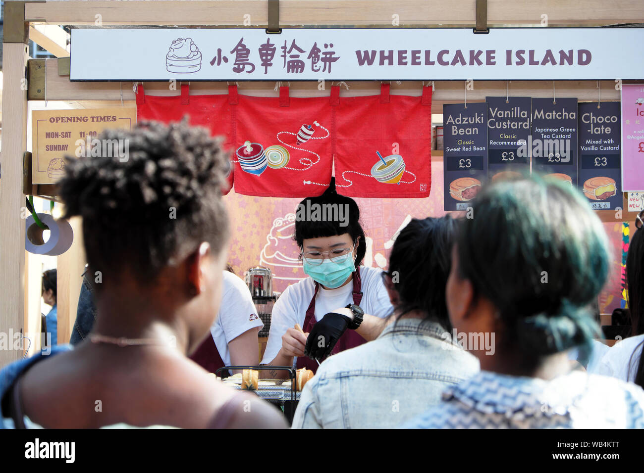 Junge Frau in hygienemaske Servieren von Speisen an Wheelcake Insel Taiwan Shop & Kunden anstehen in Spitalfields Market London England UK KATHY DEWITT Stockfoto