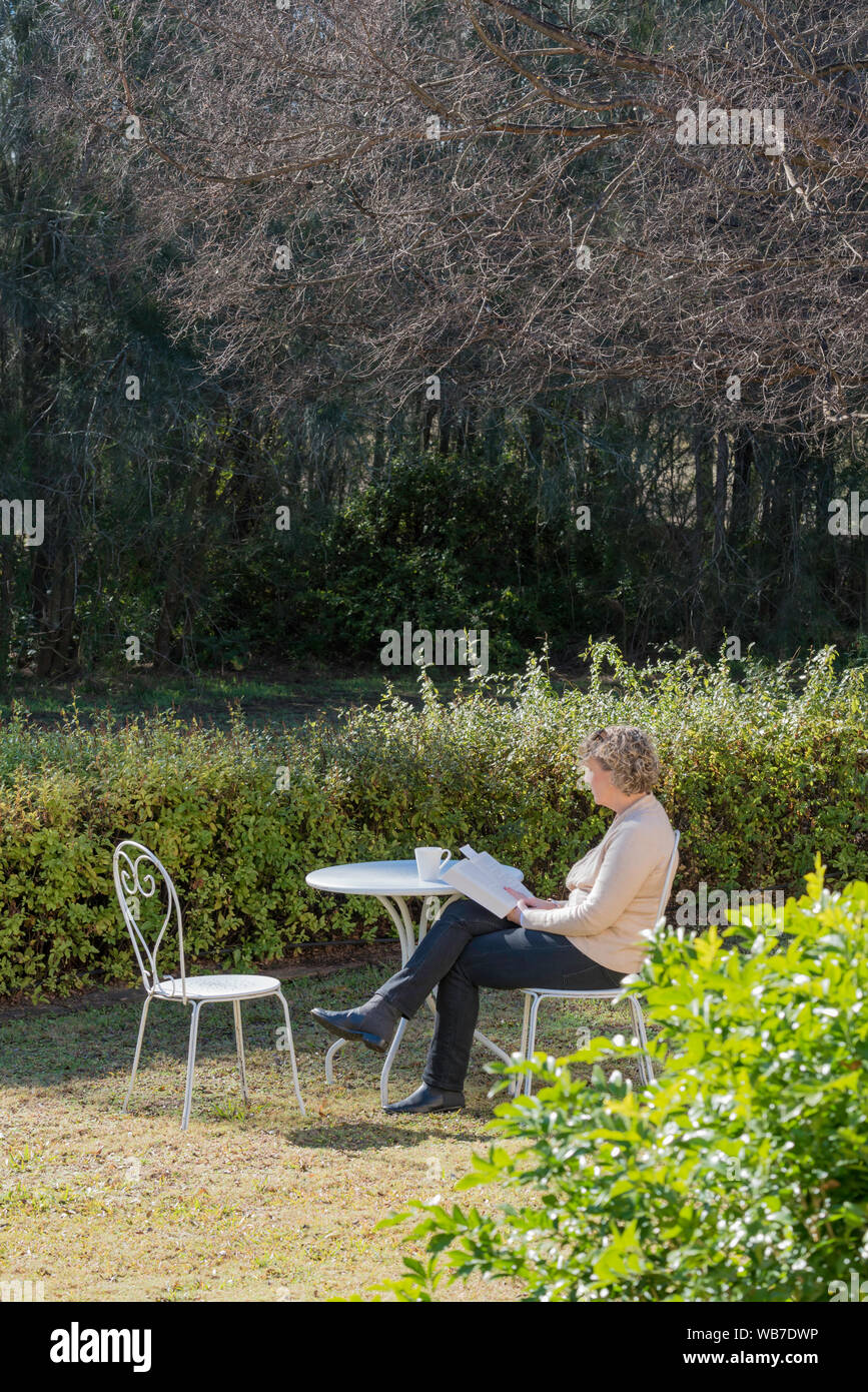 Eine weiße Frau mittleren Alters sitzt an einem weißen Tisch und Stühlen im Morgen winter Sonnenlicht im Garten mit einer Tasse Tee ein Buch lesen Stockfoto