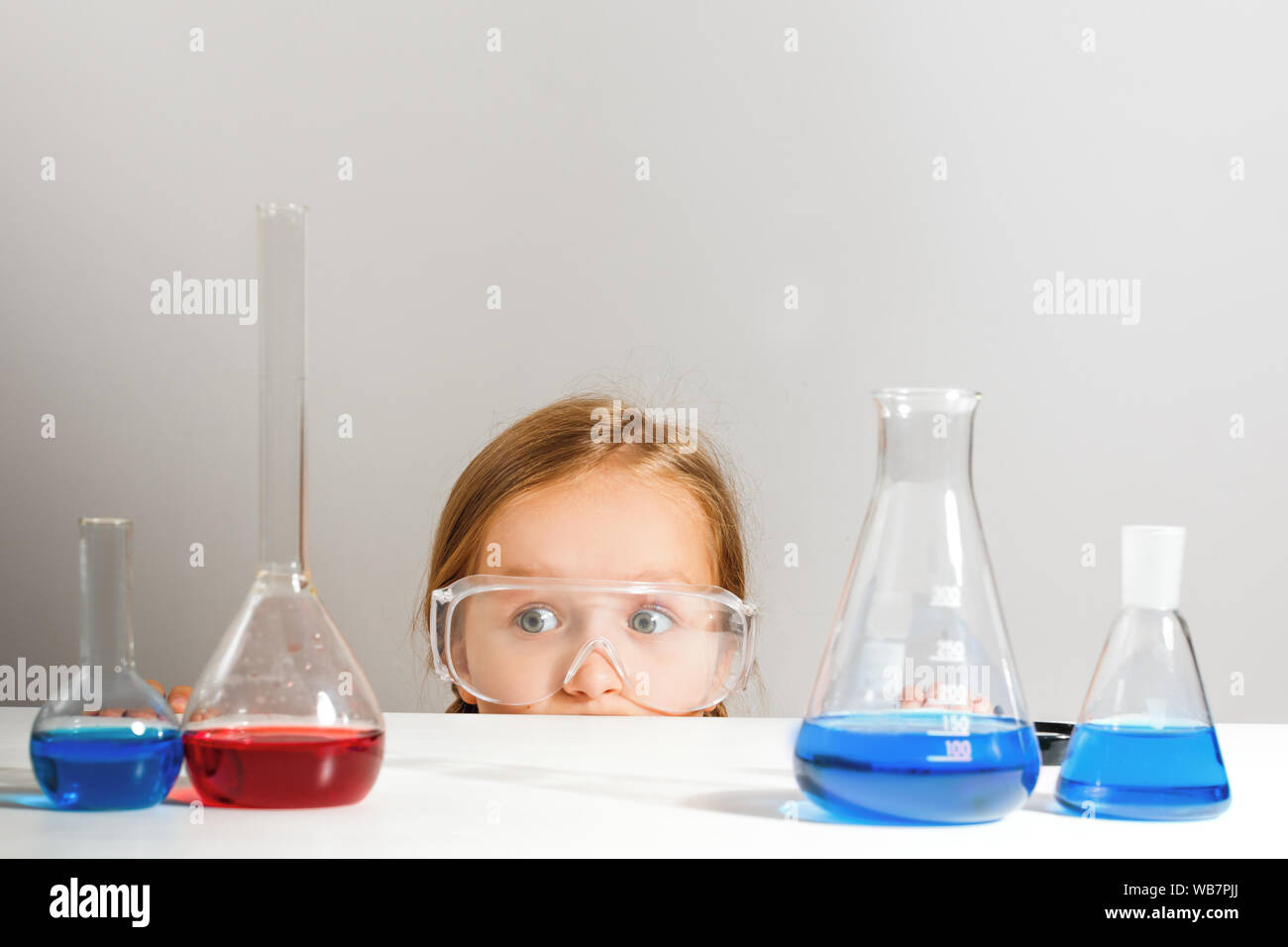 Ein kleines Mädchen in Schutzbrille versteckt sich in einem Chemielabor. Das Kind sieht aus der Tabelle. Stockfoto