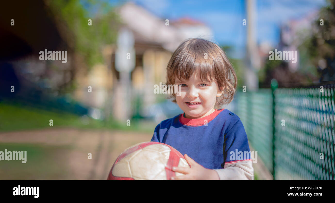 Portrait von kleinen Jungen Kind Junge Spaß spielen Fußball-Fußball-Spiel auf sonnigen Sommertag auf Kamera Holding eine schmutzige Kugel in öffentlichen Stadt u Stockfoto