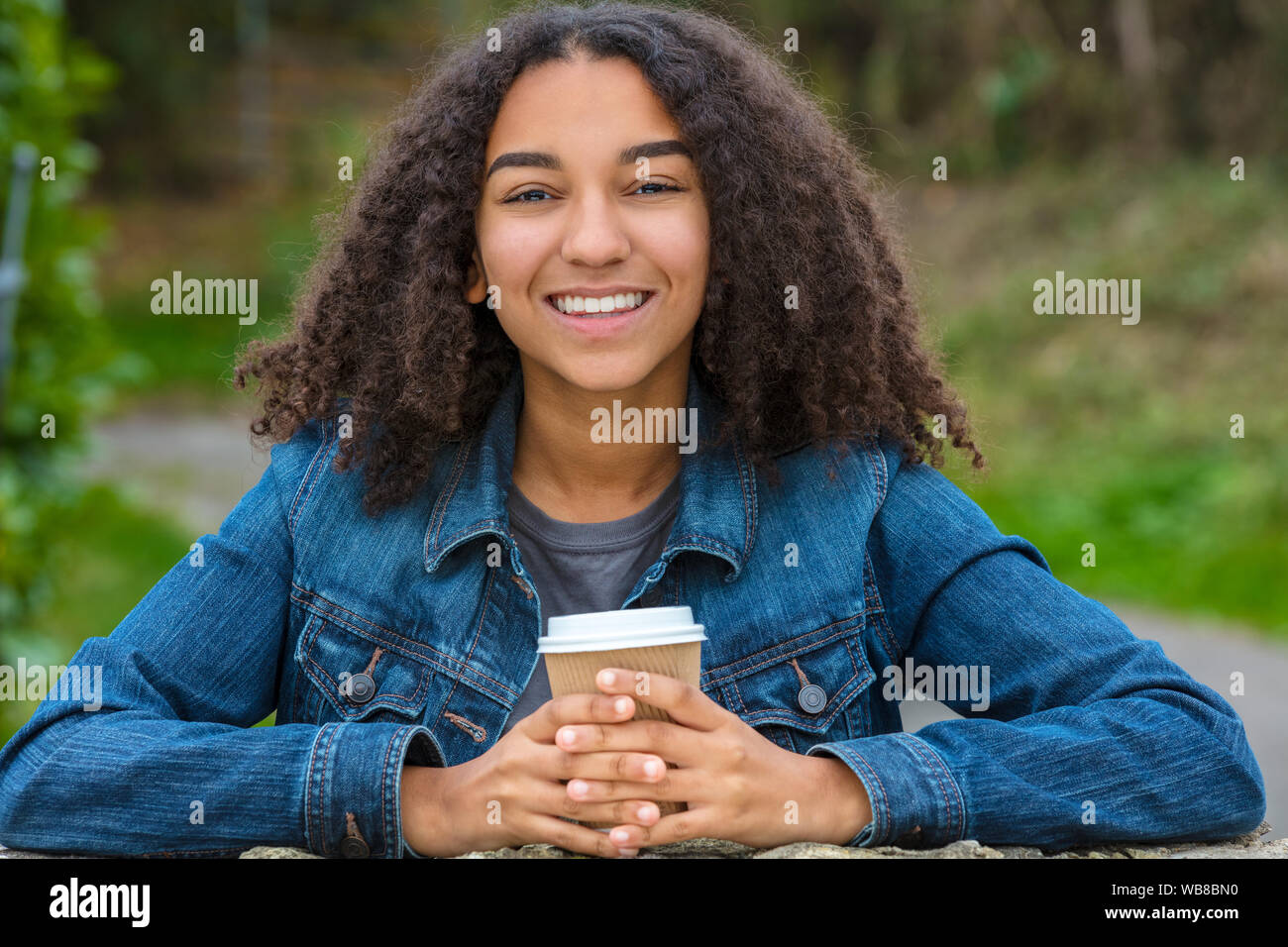Schöne happy biracial Mixed Race African American Girl Teenager weibliche junge Frau lächelnd mit perfekte Zähne trinken Kaffee zum Mitnehmen vor Verschleiß Stockfoto