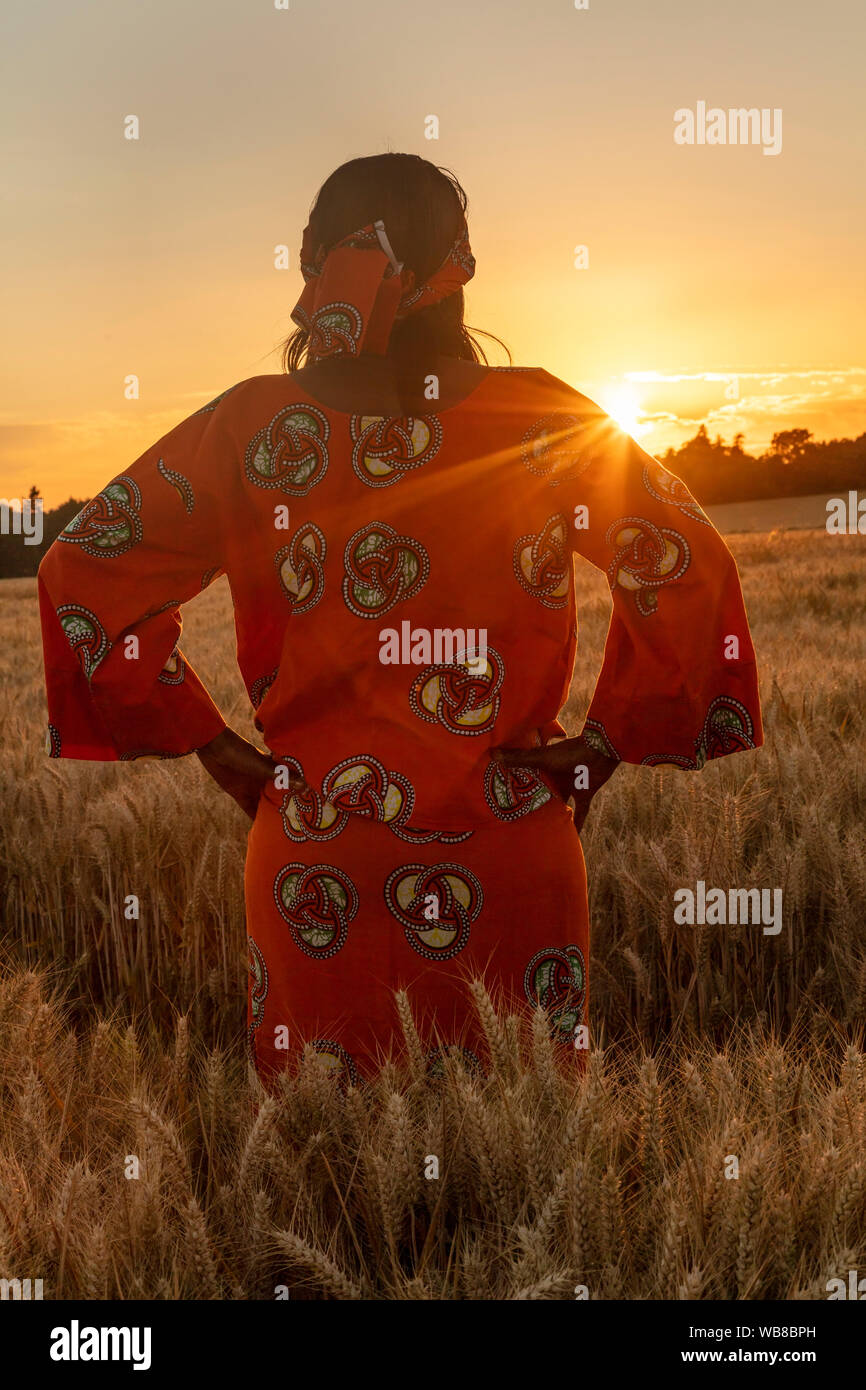 Afrikanische Frau in traditioneller Kleidung stehen, Suchen, die Hände auf den Hüften, im Bereich der Gerste oder Weizen bei Sonnenuntergang oder Sunrise Stockfoto