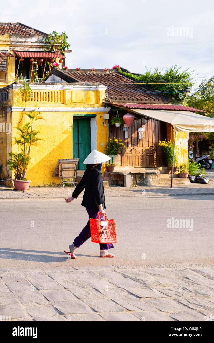 Frau mit traditionellen vietnamesischen Hut und Tasche in der Straße in der Altstadt von Hoi An in Südostasien in Vietnam am Morgen. Stadtbild mit Lady und Stockfoto