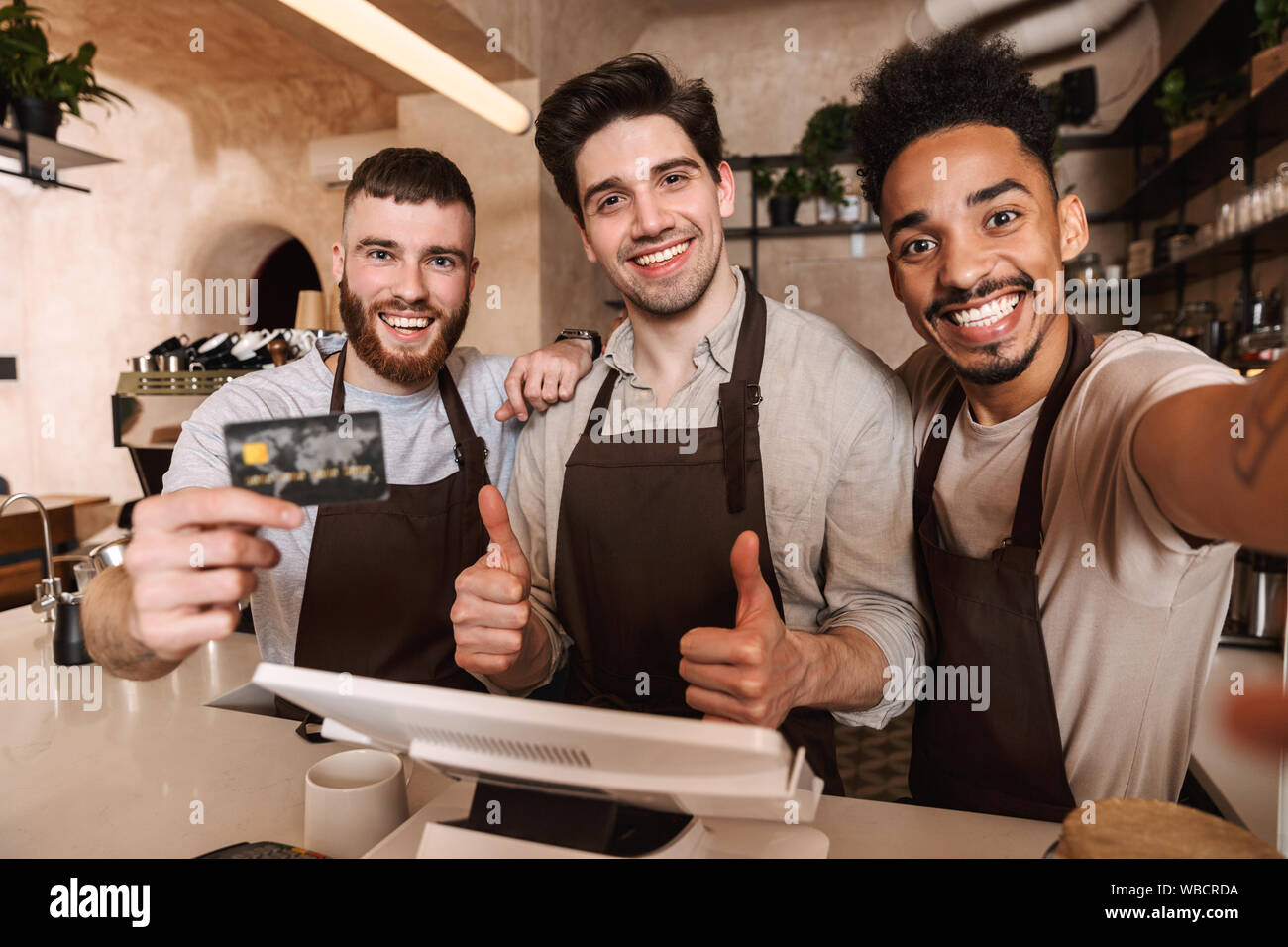 Drei fröhliche Männer baristas steht hinter der Theke im Coffee Shop, ein selfie, Kreditkarte Stockfoto