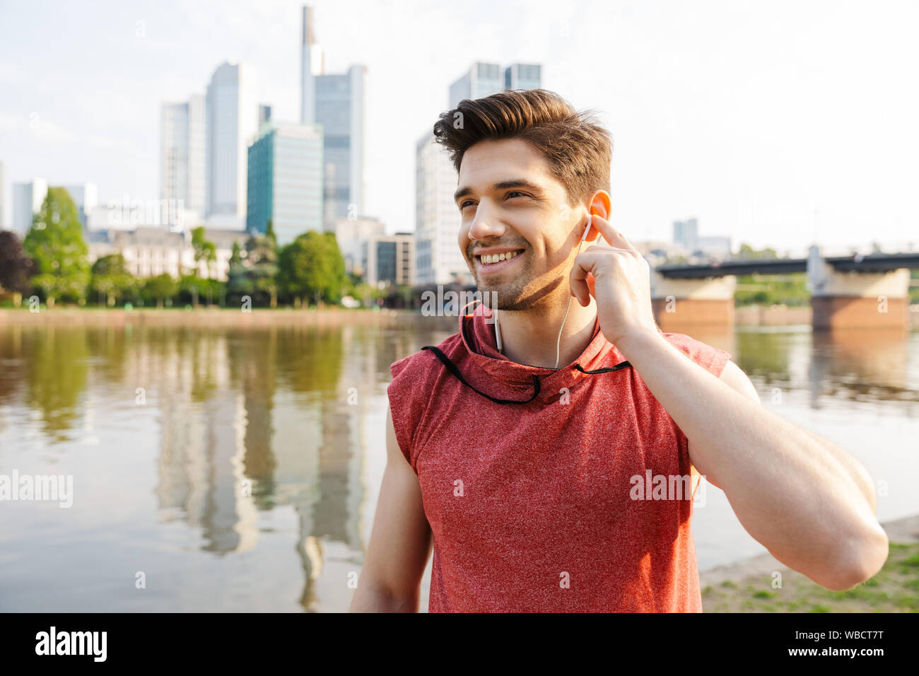 Fotos von lächelnden athletischer Mann mit Sportswear über Kopfhörer, während Sie in der Nähe von City riverfront arbeiten Stockfoto