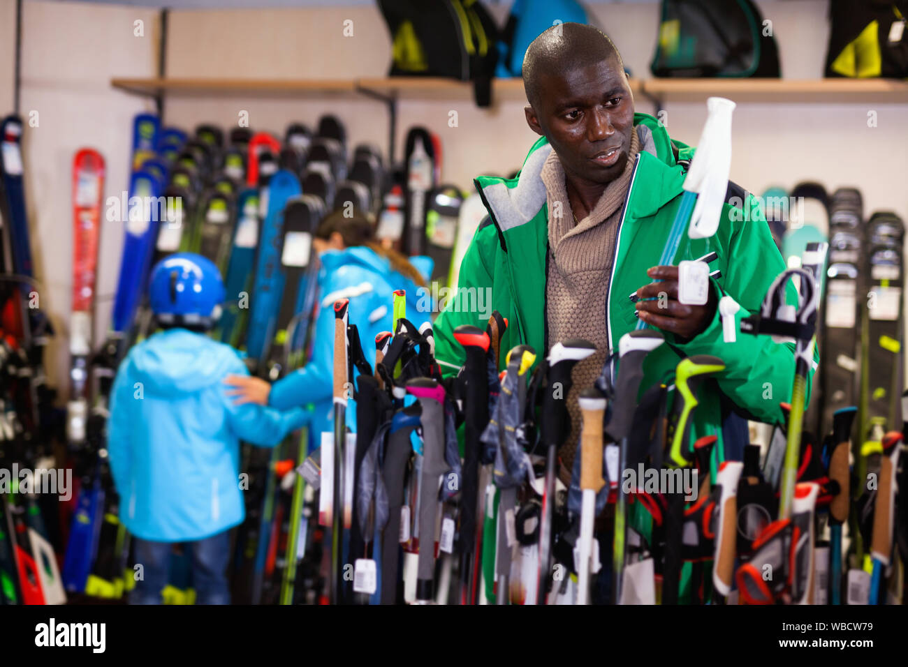 Junge fröhliche positive lächelnde African American skier Skistöcke wählen im Sport waren Store Stockfoto
