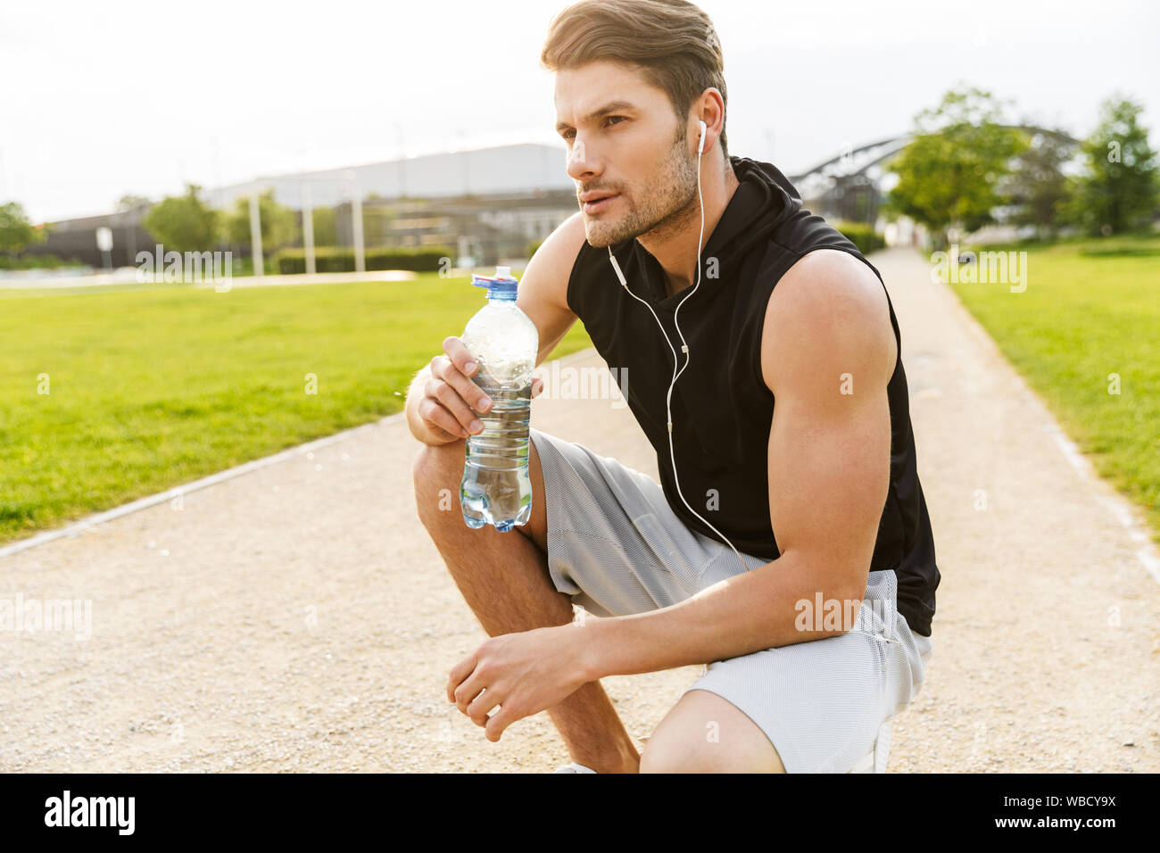Bild der männlichen Mann in Sportswear Trinkwasser während der Arbeit aus mit Ohrhörer in Green Park im Freien in Morgen Stockfoto