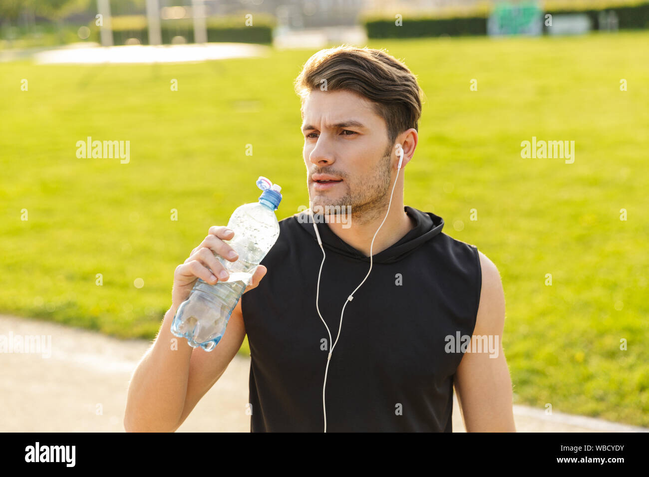 Bild der männlichen Mann in Sportswear Trinkwasser während der Arbeit aus mit Kopfhörern und läuft im Green Park im Freien in Morgen Stockfoto