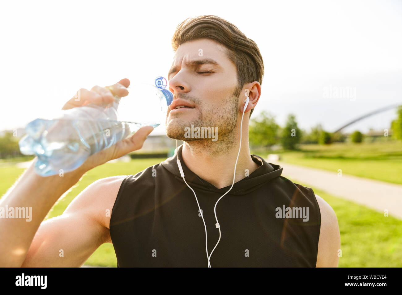 Bild der kaukasischen Mann in Sportswear Trinkwasser während der Arbeit aus mit Kopfhörern und läuft im Green Park im Freien in Morgen Stockfoto