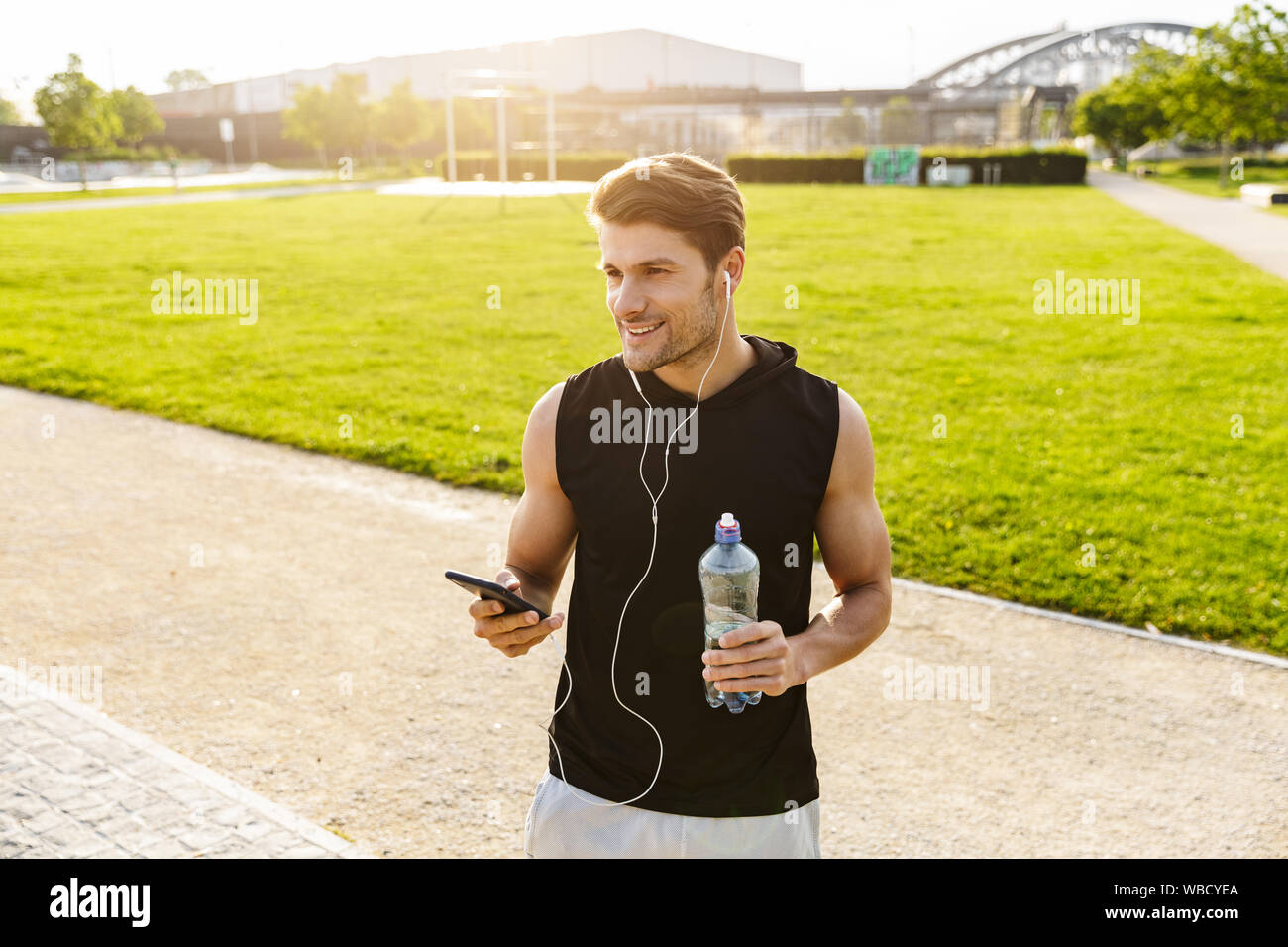 Bild der muskulösen Mann in Sportswear Trinkwasser und Holding Handy während der Ausführung mit Ohrhörer in Green Park im Freien Stockfoto