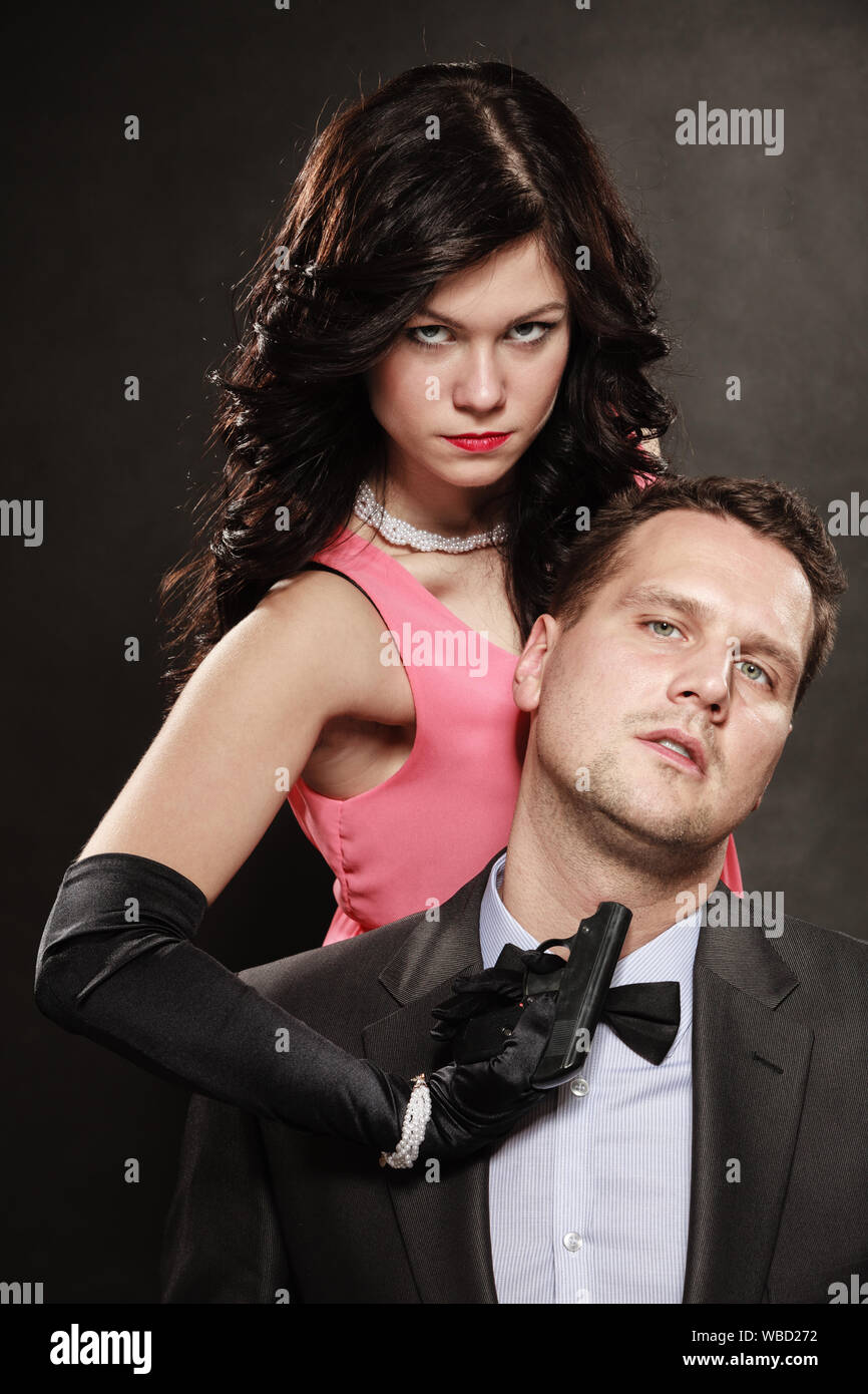 Szene der Gewalt mit schusswaffe zwischen Männern und Frauen. Elegante Dame holding Waffe gegen Sitzt Mann in Anzug auf schwarz grau Hintergrund im Studio. Stockfoto