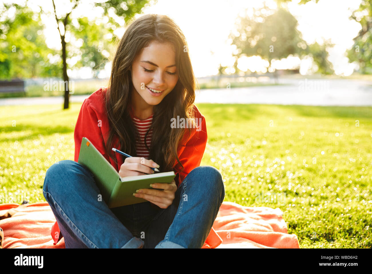 Bild der kaukasischen girl Student trägt die rote Jacke lächelnd und Verfassen von Notizen Tagebuch während auf Decke im Green Park sitzen Stockfoto