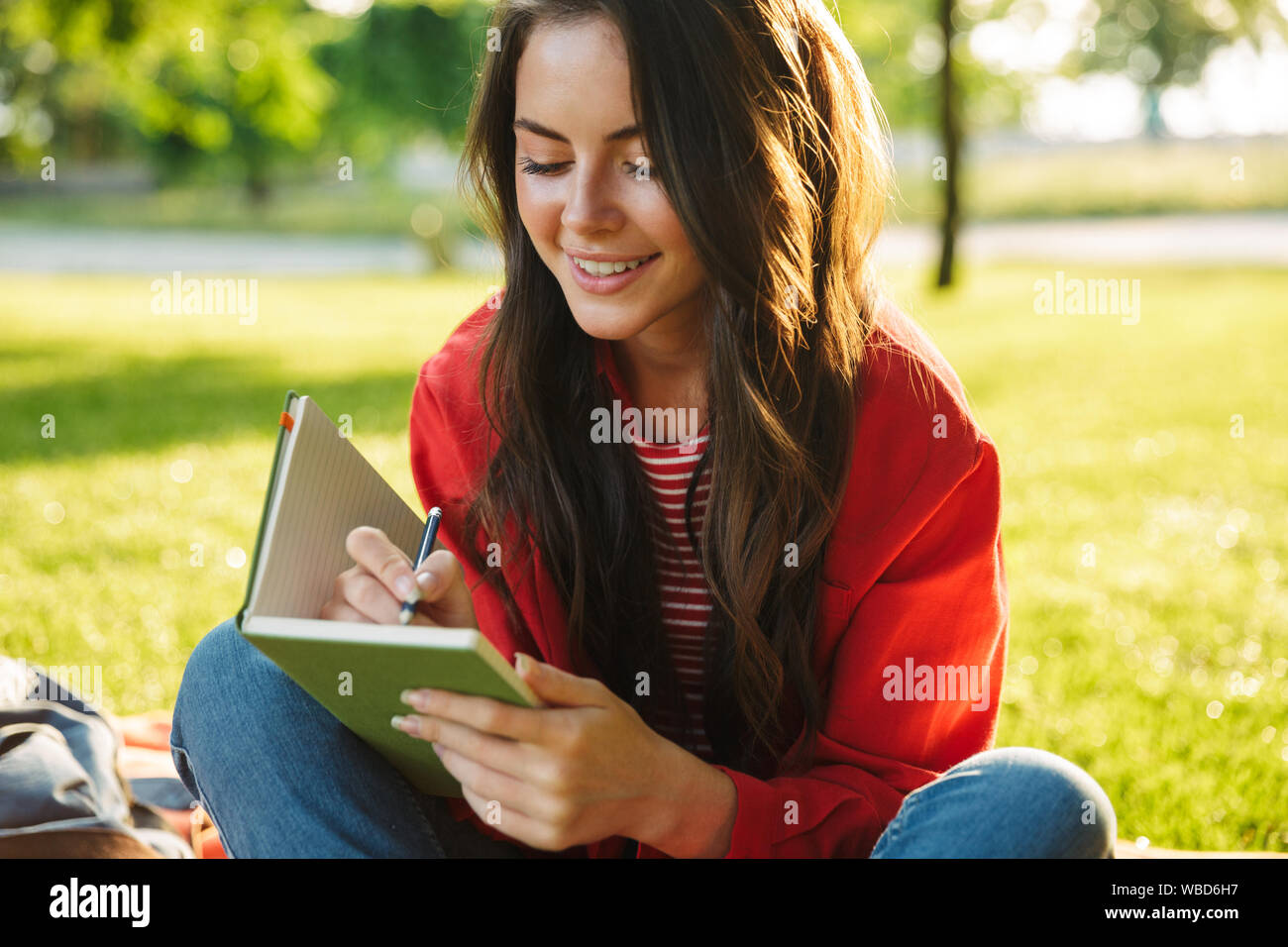 Bild der hübschen Mädchen Student trägt die rote Jacke lächelnd und Verfassen von Notizen Tagebuch während auf Decke im Green Park sitzen Stockfoto