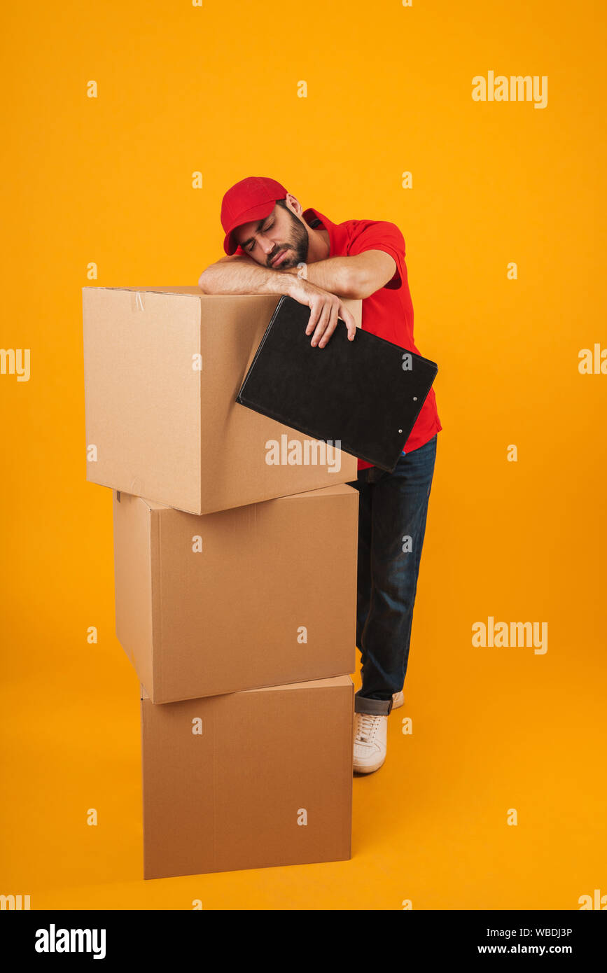 Bild von müde Lieferung Mann in der roten Uniform schlafen im Stehen mit Zwischenablage über Verpackung Kisten über Gelb Hintergrund isoliert Stockfoto