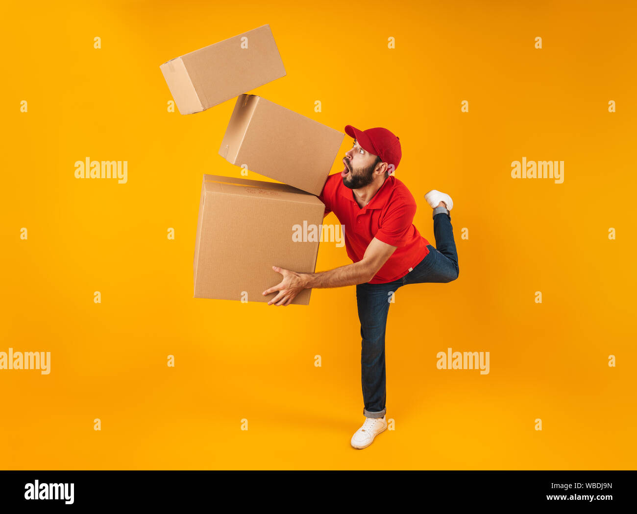 Volle Länge Portrait von schockiert Lieferung Mann in der roten Uniform tragen, die Verpackung Kisten über Gelb Hintergrund isoliert Stockfoto