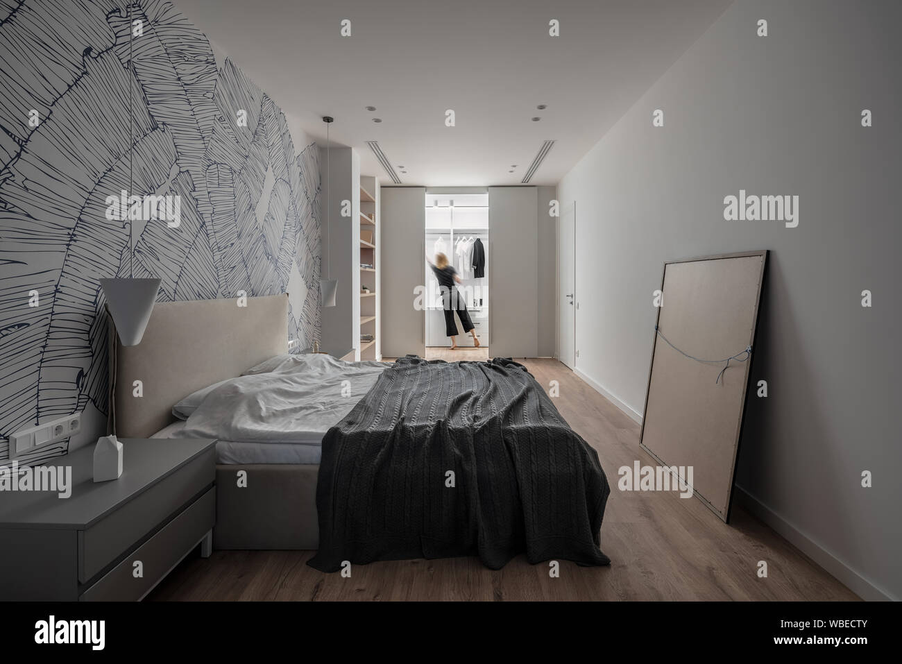 Modernes Schlafzimmer mit weißen Wänden mit Mustern und Parkett auf dem Fußboden. Es gibt ein Doppelbett, Nachttische, Holzregale, Tür, Hängeleuchte, Stockfoto