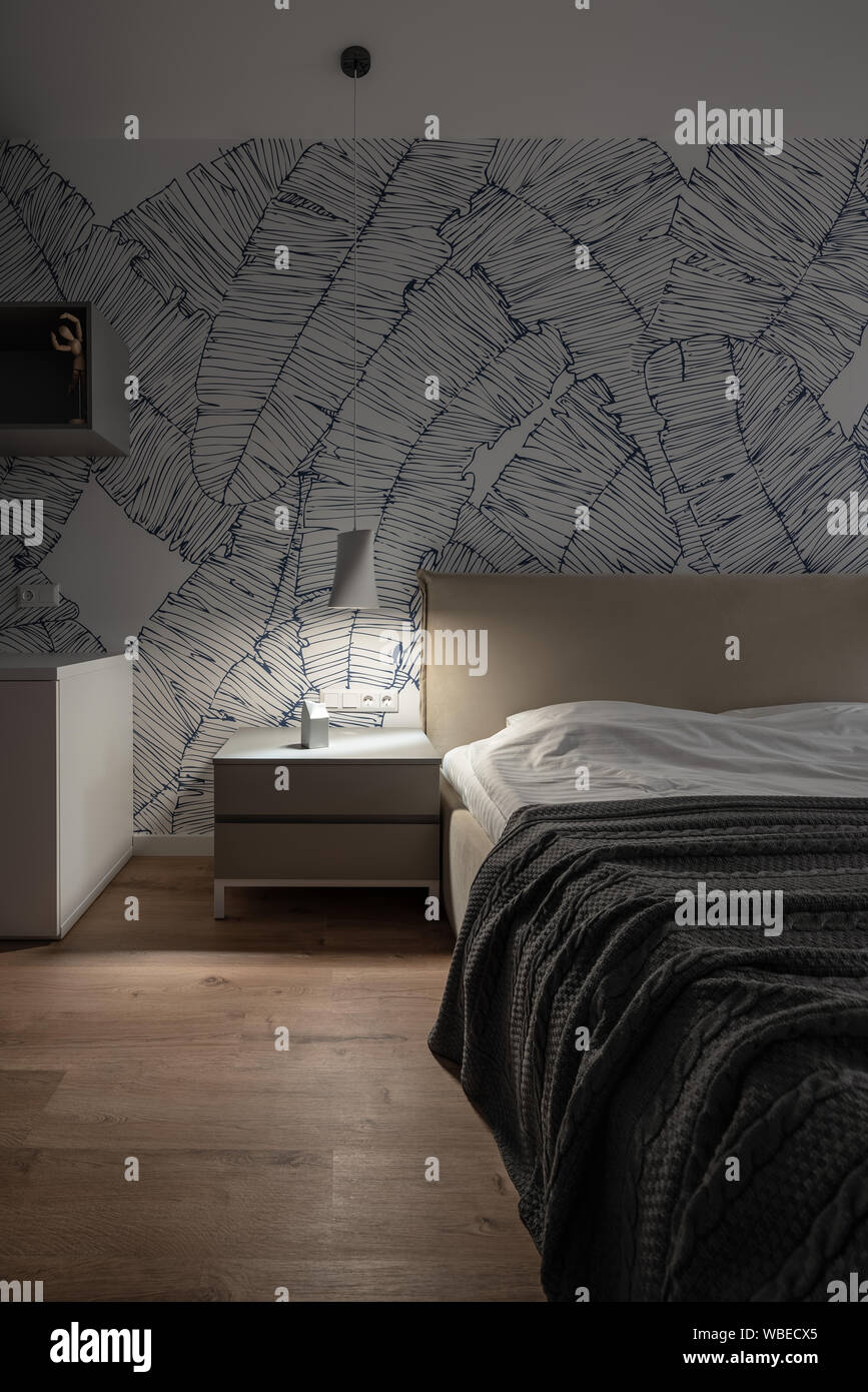 Beleuchtetes Schlafzimmer mit weißen Wand mit Mustern und Parkett auf dem Fußboden. Es gibt ein Doppelbett mit Bettwäsche und einem dunklen Bettdecke, grau Nächte Stockfoto